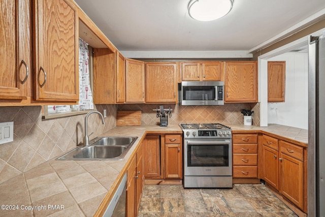 kitchen with tile countertops, decorative backsplash, appliances with stainless steel finishes, brown cabinetry, and a sink