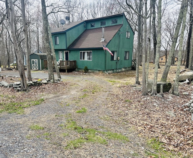view of front facade with a wooden deck, a shed, a shingled roof, dirt driveway, and an outbuilding
