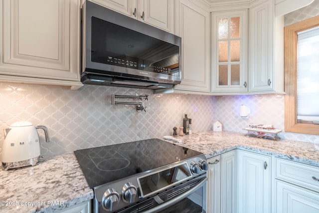 kitchen with glass insert cabinets, light stone countertops, backsplash, and appliances with stainless steel finishes