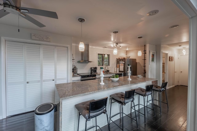 kitchen with appliances with stainless steel finishes, wall chimney range hood, a ceiling fan, dark wood-style floors, and a kitchen breakfast bar