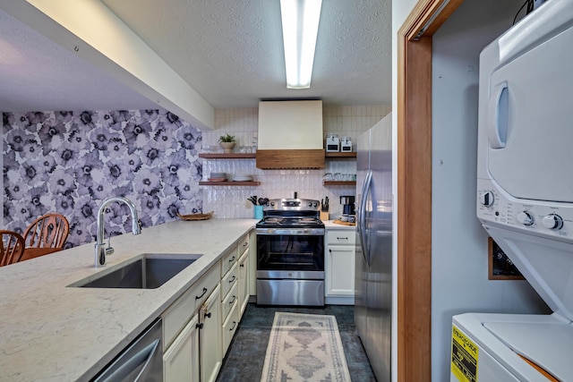 kitchen with stacked washing maching and dryer, open shelves, a sink, appliances with stainless steel finishes, and wall chimney exhaust hood