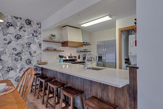 kitchen with open shelves, wall chimney range hood, stacked washer and dryer, stainless steel appliances, and a sink