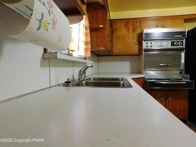 kitchen with brown cabinetry, open shelves, a sink, light countertops, and wall oven