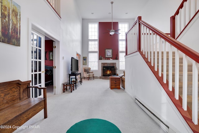 interior space with a warm lit fireplace, a baseboard radiator, a high ceiling, a ceiling fan, and stairs