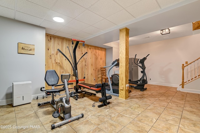 workout area featuring a paneled ceiling, tile patterned flooring, wooden walls, and baseboards