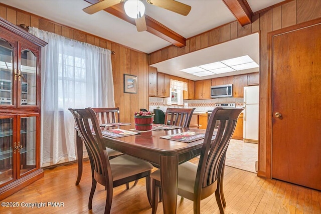 dining room with ceiling fan, wooden walls, beam ceiling, and light hardwood / wood-style flooring