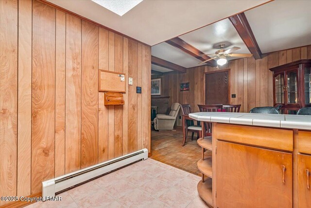 kitchen with ceiling fan, a baseboard radiator, tile countertops, beamed ceiling, and wood walls