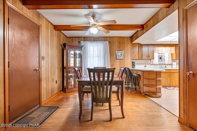 dining space with ceiling fan, light wood-type flooring, beamed ceiling, and wood walls