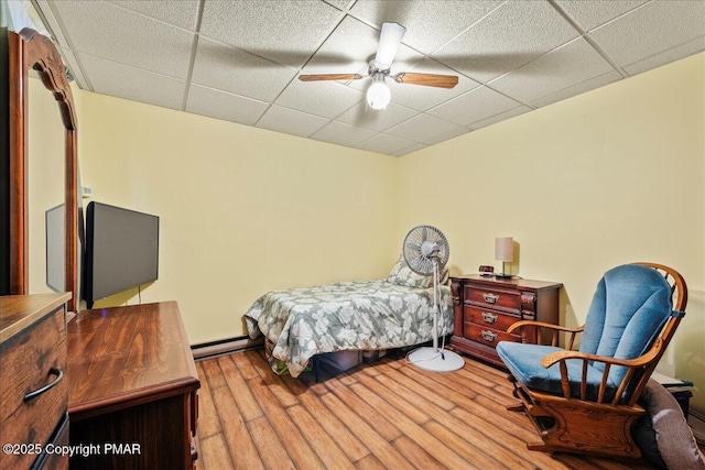 bedroom featuring a drop ceiling, wood finished floors, a baseboard radiator, and a ceiling fan