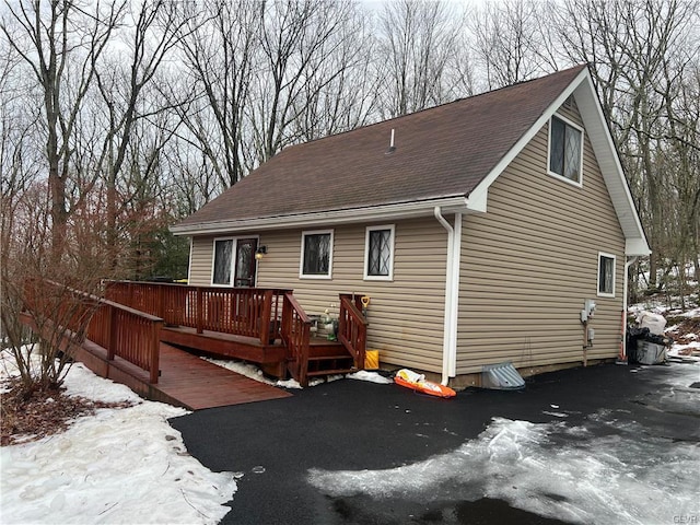 snow covered back of property featuring a shingled roof, driveway, and a wooden deck