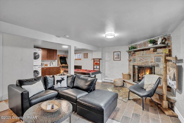 living room featuring light wood-type flooring, billiards, stacked washing maching and dryer, a stone fireplace, and a baseboard radiator