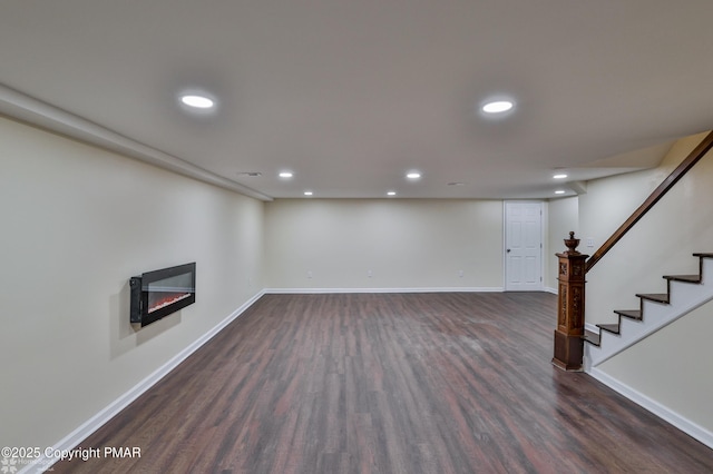 finished below grade area featuring a glass covered fireplace, dark wood finished floors, recessed lighting, baseboards, and stairs