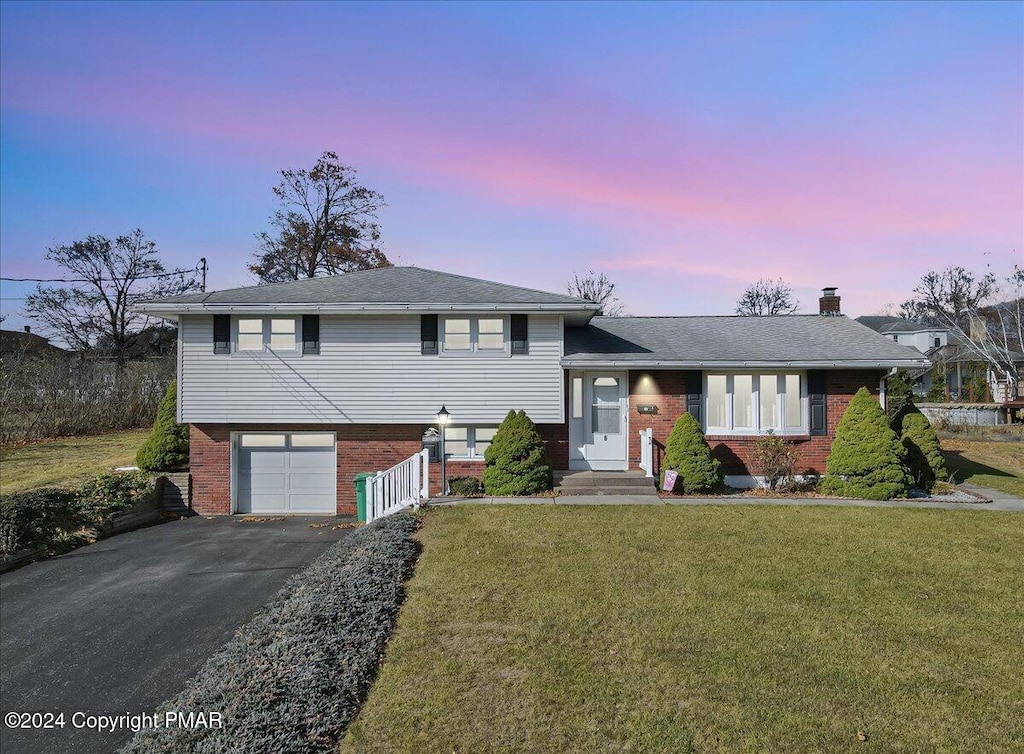 split level home featuring brick siding, aphalt driveway, a chimney, a yard, and an attached garage