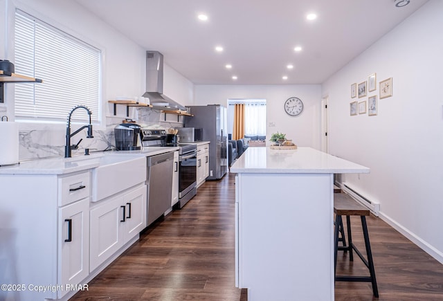 kitchen with wall chimney exhaust hood, appliances with stainless steel finishes, a baseboard heating unit, a kitchen island, and a kitchen breakfast bar