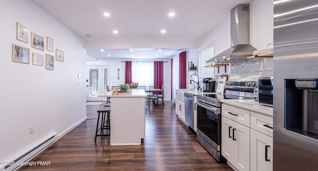 kitchen with a baseboard heating unit, stainless steel appliances, white cabinets, wall chimney range hood, and a kitchen bar