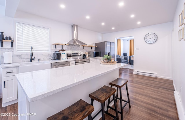 kitchen with a baseboard radiator, decorative backsplash, appliances with stainless steel finishes, a sink, and wall chimney range hood