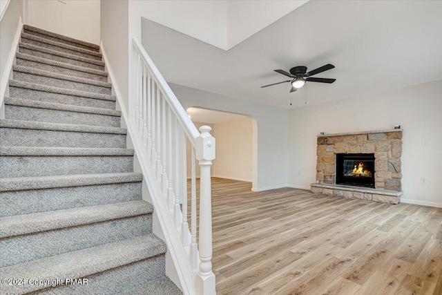 staircase with baseboards, arched walkways, a ceiling fan, wood finished floors, and a stone fireplace