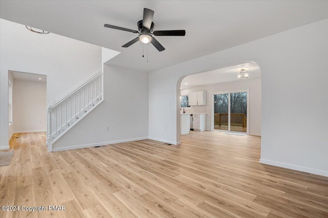 unfurnished living room with arched walkways, light wood-style flooring, ceiling fan, baseboards, and stairs