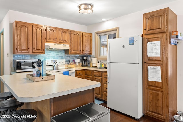 kitchen with under cabinet range hood, white appliances, a kitchen breakfast bar, light countertops, and decorative backsplash