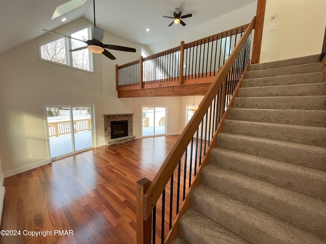 stairs featuring ceiling fan, a stone fireplace, wood finished floors, high vaulted ceiling, and baseboards