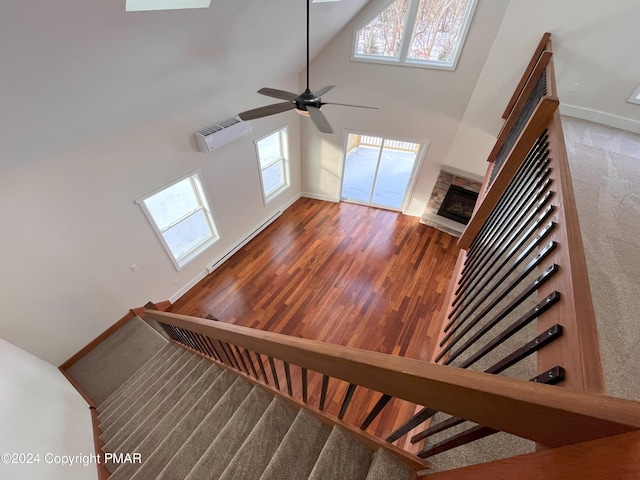 stairway with a wall unit AC, a baseboard heating unit, a stone fireplace, wood finished floors, and high vaulted ceiling