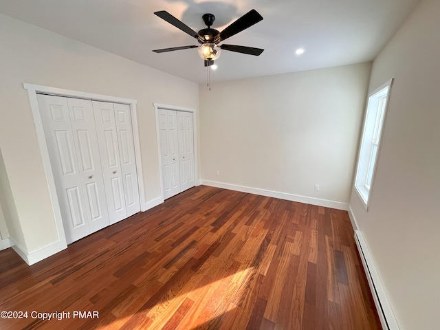 unfurnished bedroom featuring a baseboard heating unit, baseboards, multiple closets, and dark wood-style floors