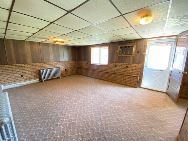 below grade area featuring carpet flooring, a drop ceiling, a wall unit AC, and radiator heating unit