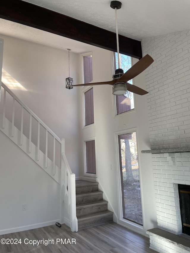 stairway featuring a brick fireplace, beamed ceiling, a high ceiling, wood finished floors, and a ceiling fan