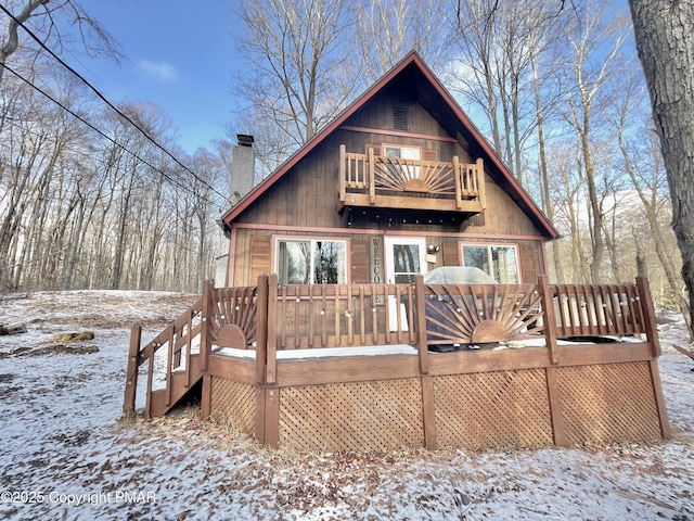 snow covered back of property featuring a balcony, a chimney, and a wooden deck