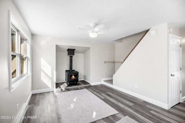 unfurnished living room with ceiling fan, wood finished floors, baseboards, stairway, and a wood stove