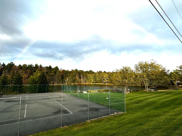 view of tennis court featuring a water view, fence, a view of trees, and a yard