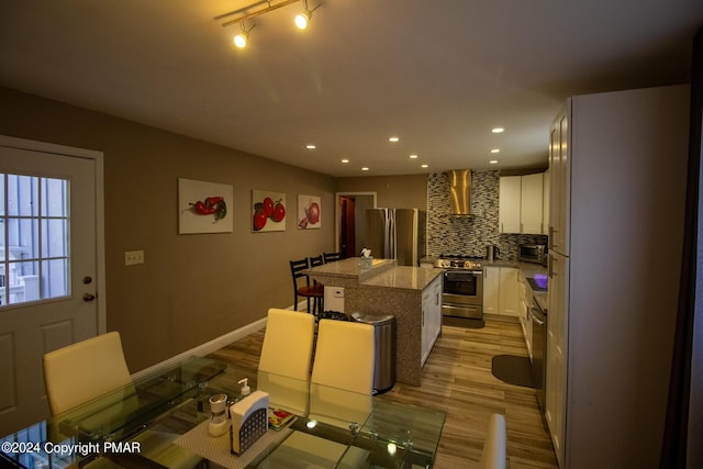 kitchen featuring light wood finished floors, tasteful backsplash, a kitchen island, stainless steel appliances, and wall chimney range hood