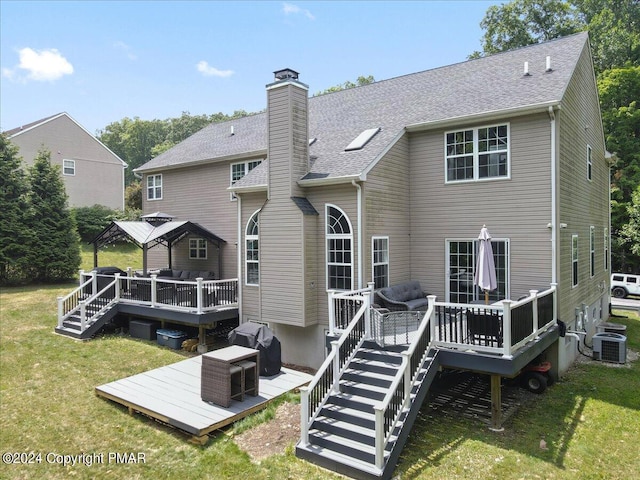 back of property with a wooden deck, a shingled roof, a chimney, central air condition unit, and a lawn