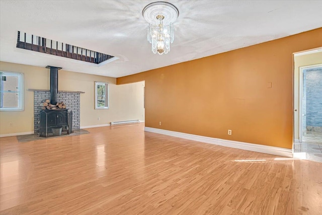 unfurnished living room featuring baseboard heating, a wood stove, light wood finished floors, a chandelier, and baseboards