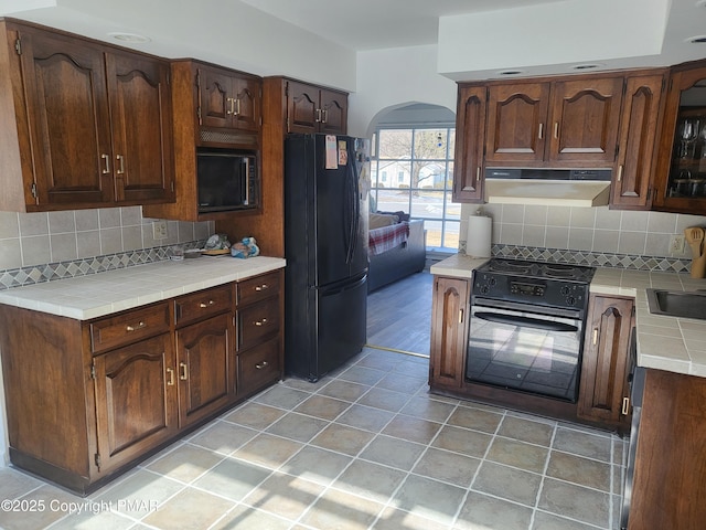 kitchen featuring arched walkways, tile countertops, under cabinet range hood, decorative backsplash, and black appliances