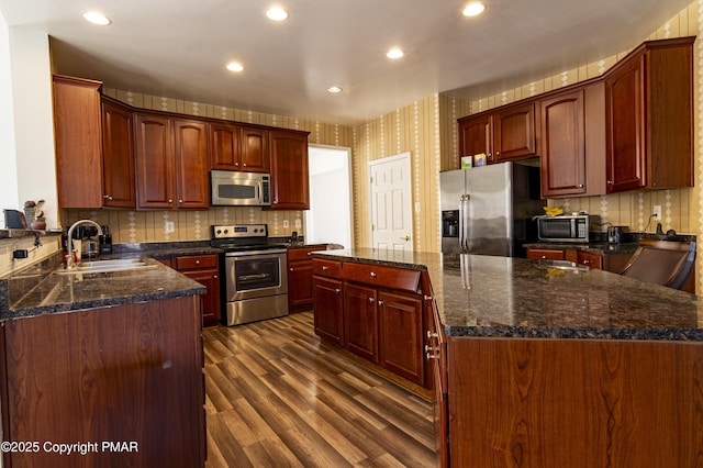 kitchen with dark wood-style flooring, a kitchen island, a sink, appliances with stainless steel finishes, and wallpapered walls