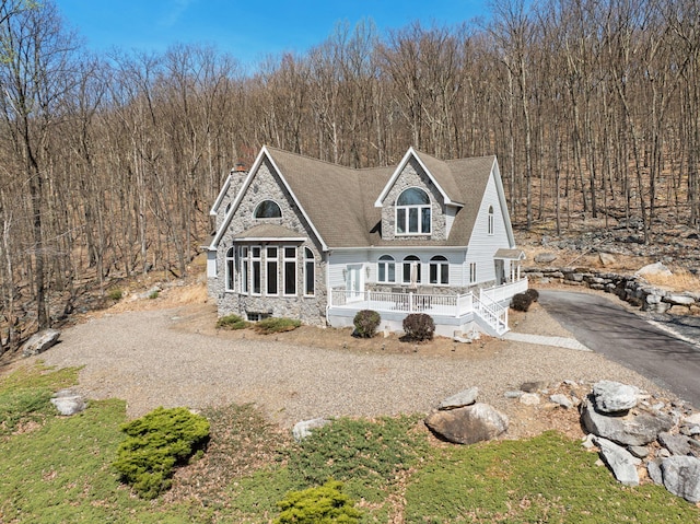 view of front facade with a porch, a forest view, driveway, a shingled roof, and stone siding