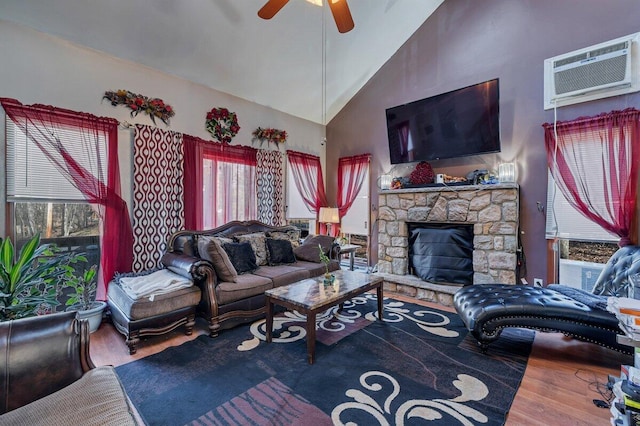 living area with high vaulted ceiling, wood finished floors, a wall unit AC, a stone fireplace, and ceiling fan