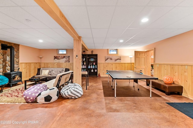 recreation room with wainscoting, a wood stove, a paneled ceiling, wood walls, and concrete floors