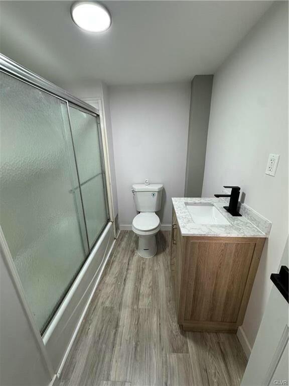 bathroom featuring toilet, vanity, wood finished floors, bath / shower combo with glass door, and baseboards