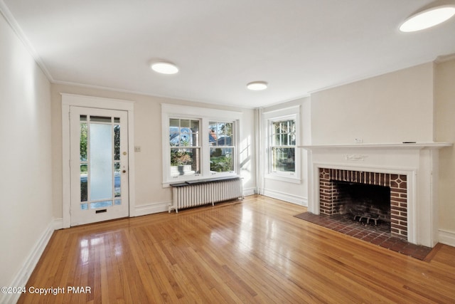 unfurnished living room featuring radiator, wood-type flooring, ornamental molding, a brick fireplace, and baseboards