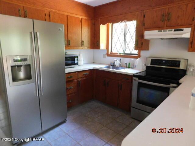 kitchen featuring a sink, stainless steel appliances, light countertops, under cabinet range hood, and brown cabinets