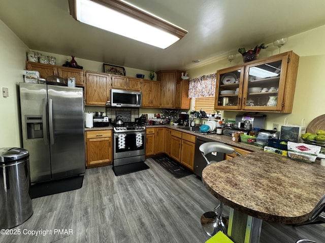 kitchen featuring brown cabinetry, glass insert cabinets, appliances with stainless steel finishes, wood finished floors, and a peninsula