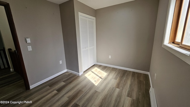 unfurnished bedroom with a closet, baseboard heating, dark wood finished floors, and baseboards