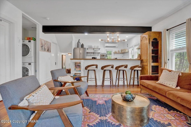 living room with wood finished floors, an AC wall unit, beam ceiling, stacked washer and clothes dryer, and an inviting chandelier