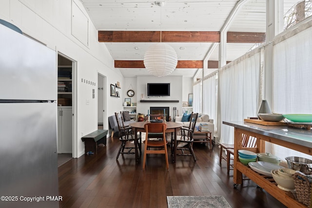 dining area featuring a warm lit fireplace, dark wood-type flooring, and beamed ceiling