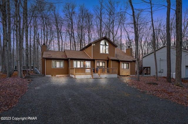 rustic home with a porch, gravel driveway, roof with shingles, and a chimney