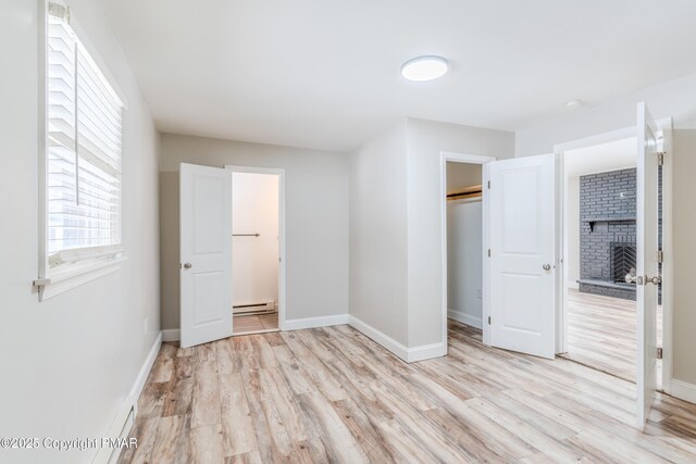 unfurnished bedroom with a spacious closet, a brick fireplace, a baseboard heating unit, light wood-type flooring, and a closet