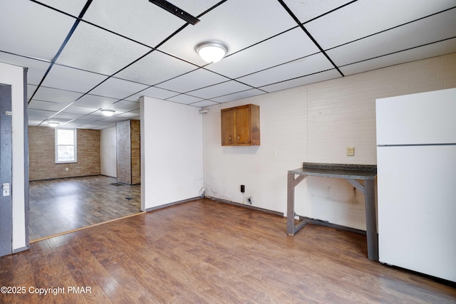 basement featuring brick wall, a drop ceiling, wood finished floors, and freestanding refrigerator