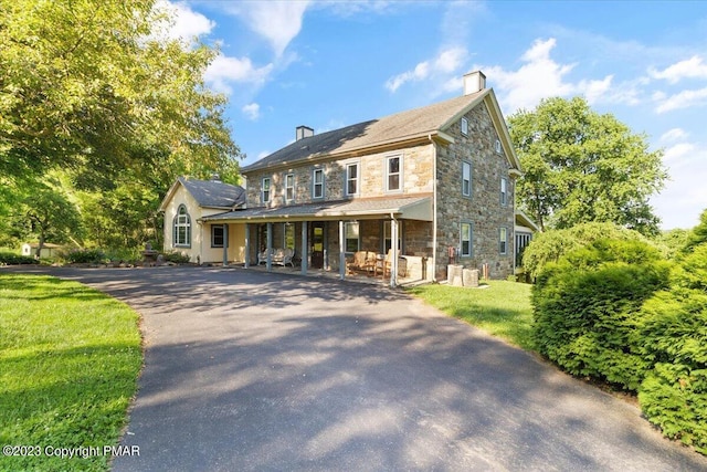 colonial-style house with aphalt driveway, a chimney, a porch, a front yard, and stone siding